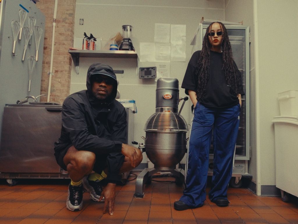 Two people, a man in a black hooded jacket and a woman with dreadlocks, standing in a room with various kitchen appliances and tools.
