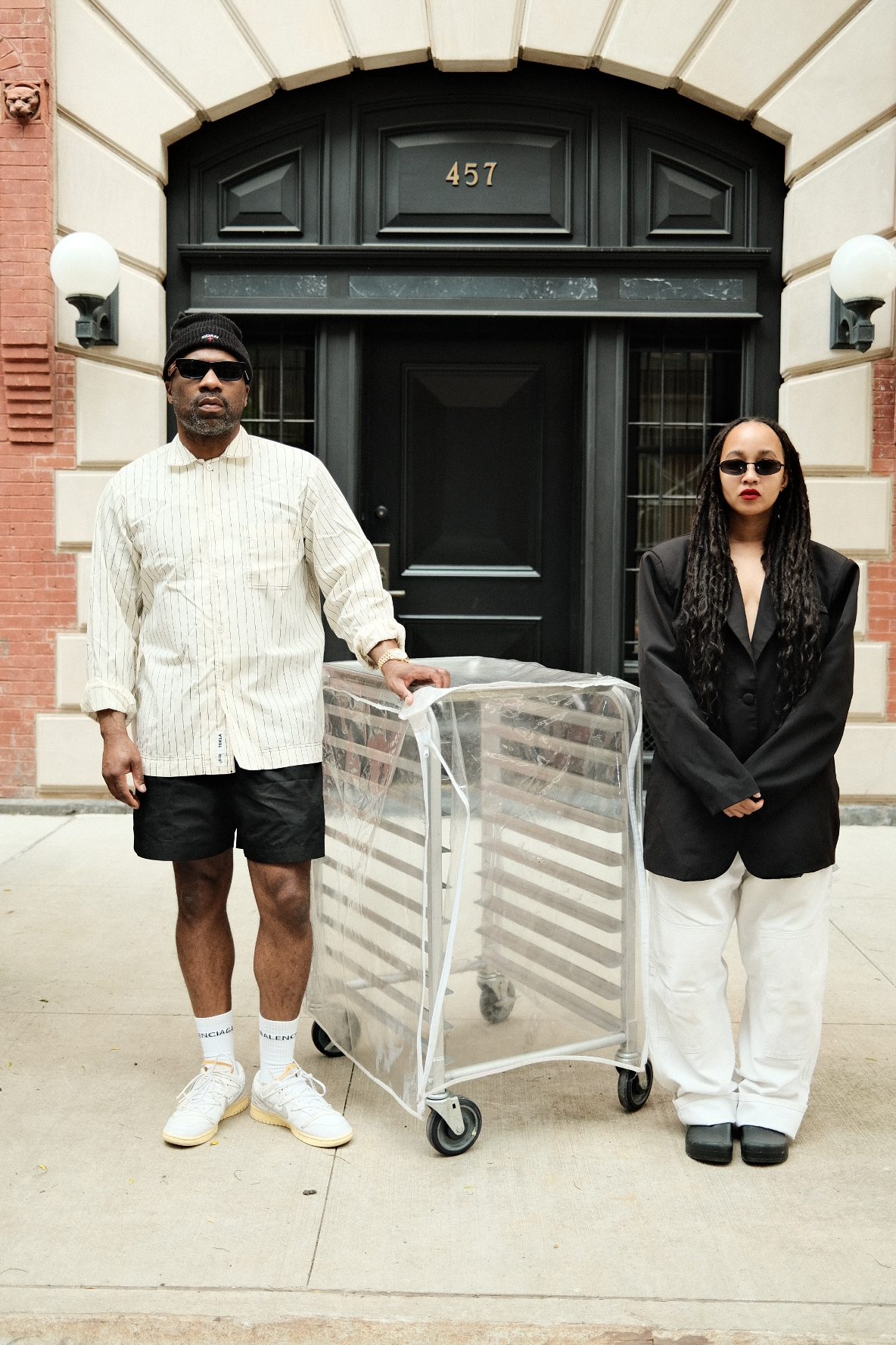Two people, a man in a white striped shirt and a woman in a black suit, standing in front of a building with the address "457" displayed above the entrance.