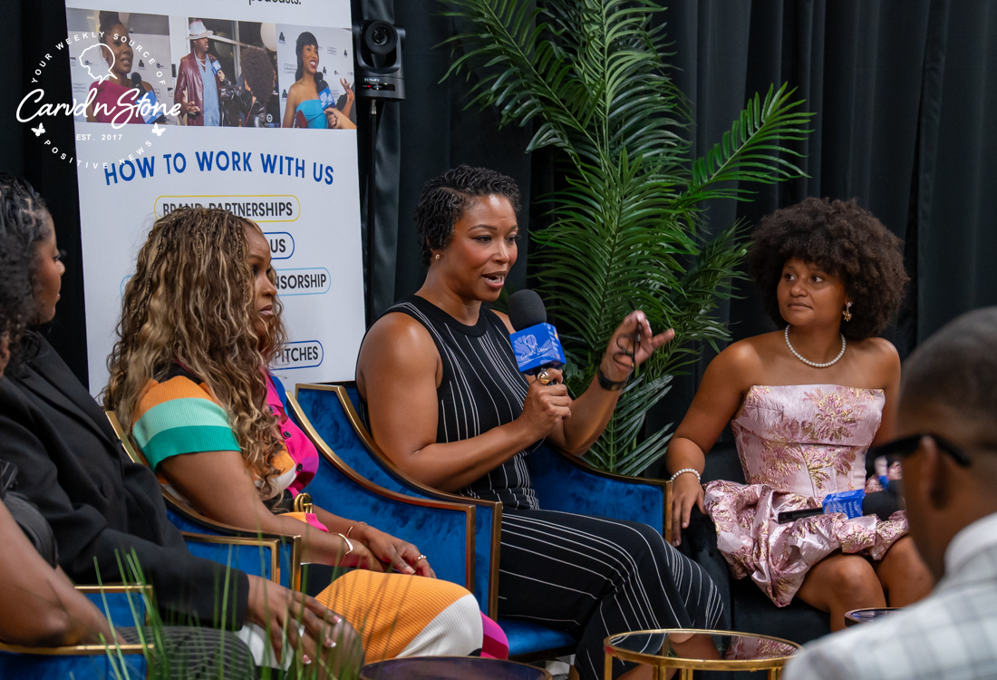A group of women, including one wearing a pink dress and another in a black and white striped outfit, sitting on chairs and discussing something, with a "How to Work with Us" sign visible in the background.