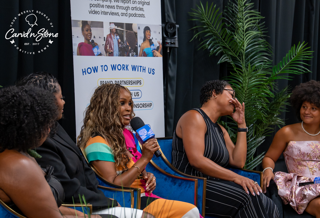 A group of people, including a woman with a microphone interviewing another woman, at an event with a "How to Work with Us" banner and palm fronds in the background.