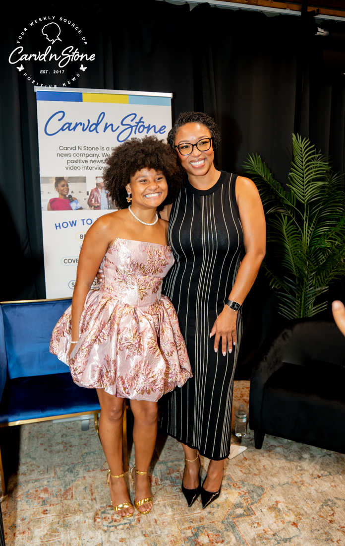 Two women standing in front of a Carvd N Stone news company backdrop, one wearing a pink floral dress and the other a black and white striped dress.