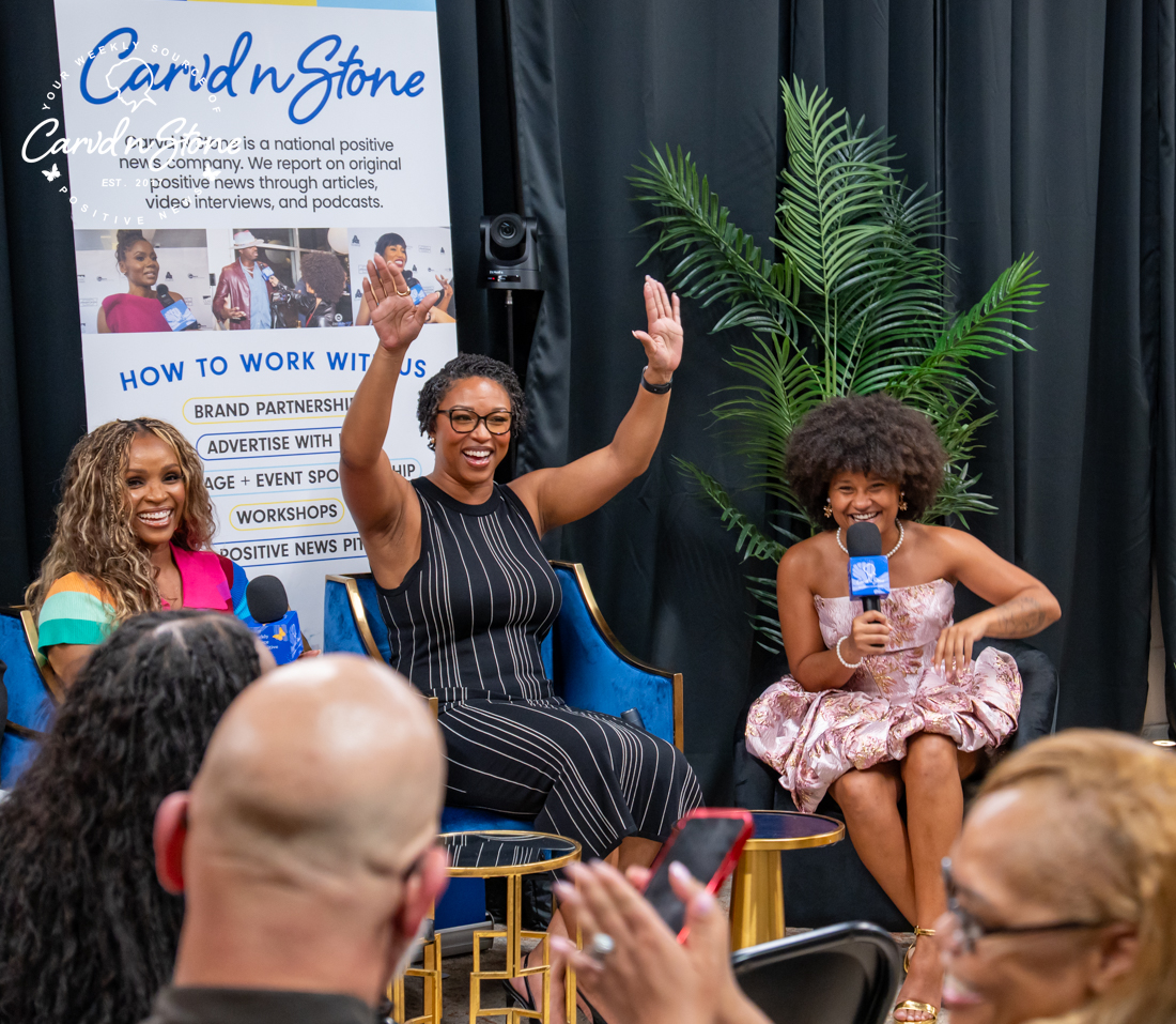 Three women on stage at a Cardi n Stone event, one speaking into a microphone while the others gesture enthusiastically. Behind them are tropical plants and a Cardi n Stone banner.