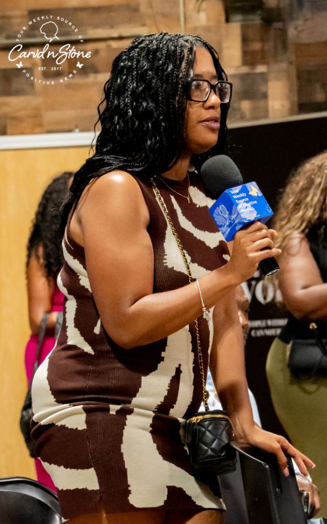 A person wearing glasses and holding a microphone, standing in front of a brick wall with a logo for "Carvin' Stone" visible.