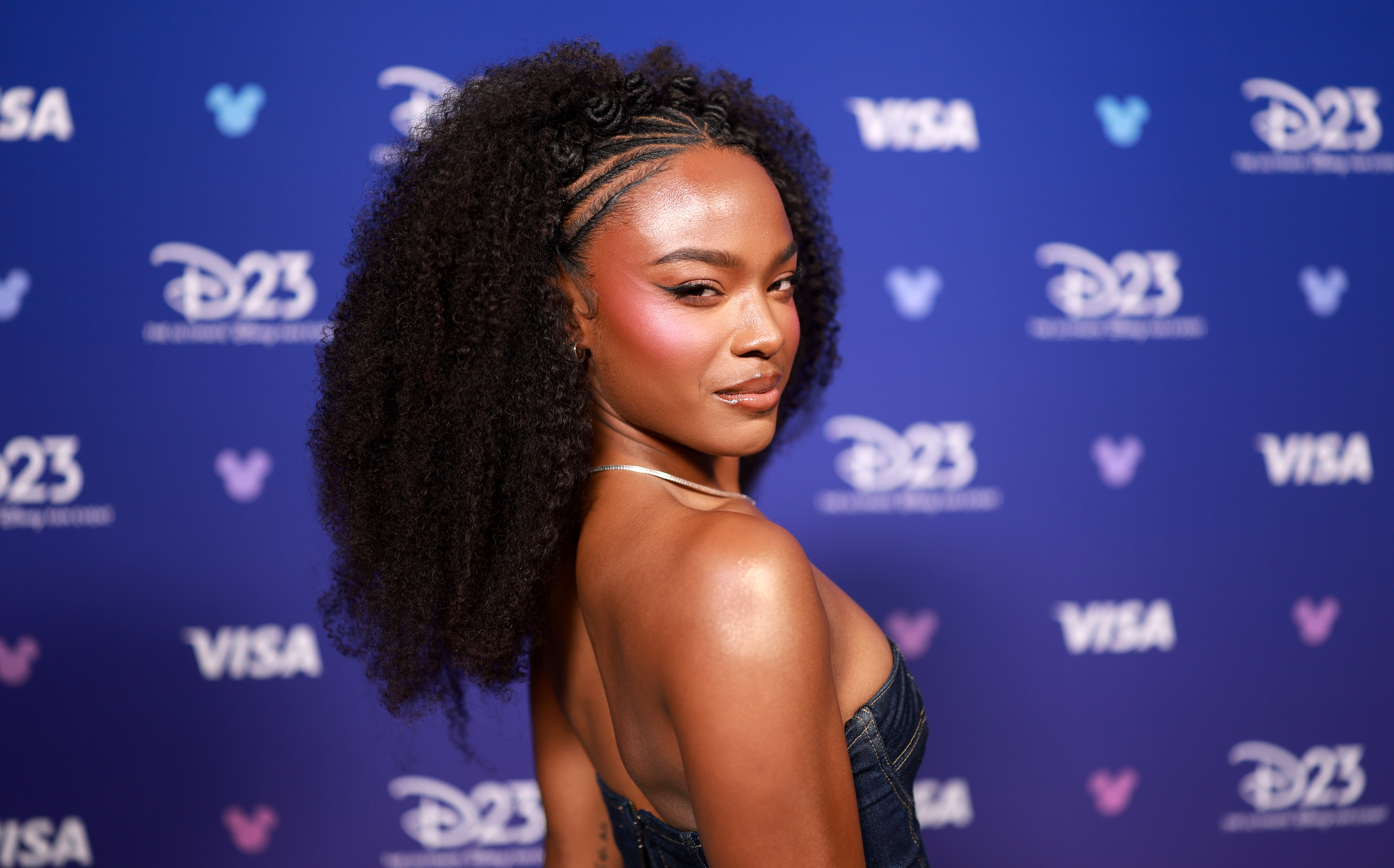 A woman with curly dark hair wearing a black halter top poses in front of a blue backdrop with Disney logos.