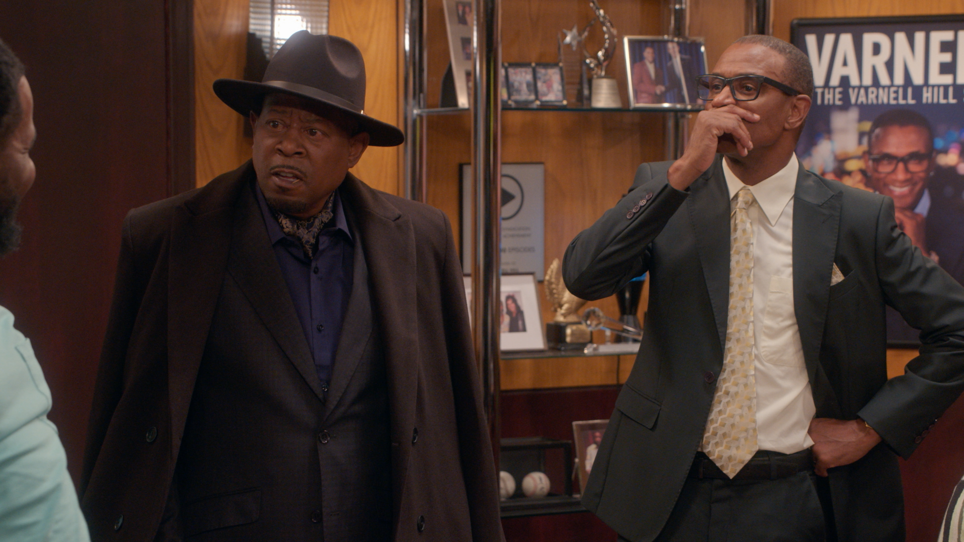 Two African American men in suits, one wearing a cowboy hat, standing in an office with framed photos and a "Varnell" sign on the wall.