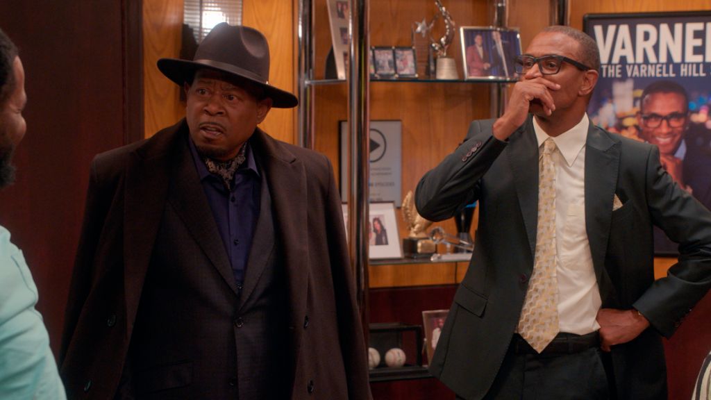 Two African American men in suits, one wearing a cowboy hat, standing in an office with framed photos and a "Varnell" sign on the wall.