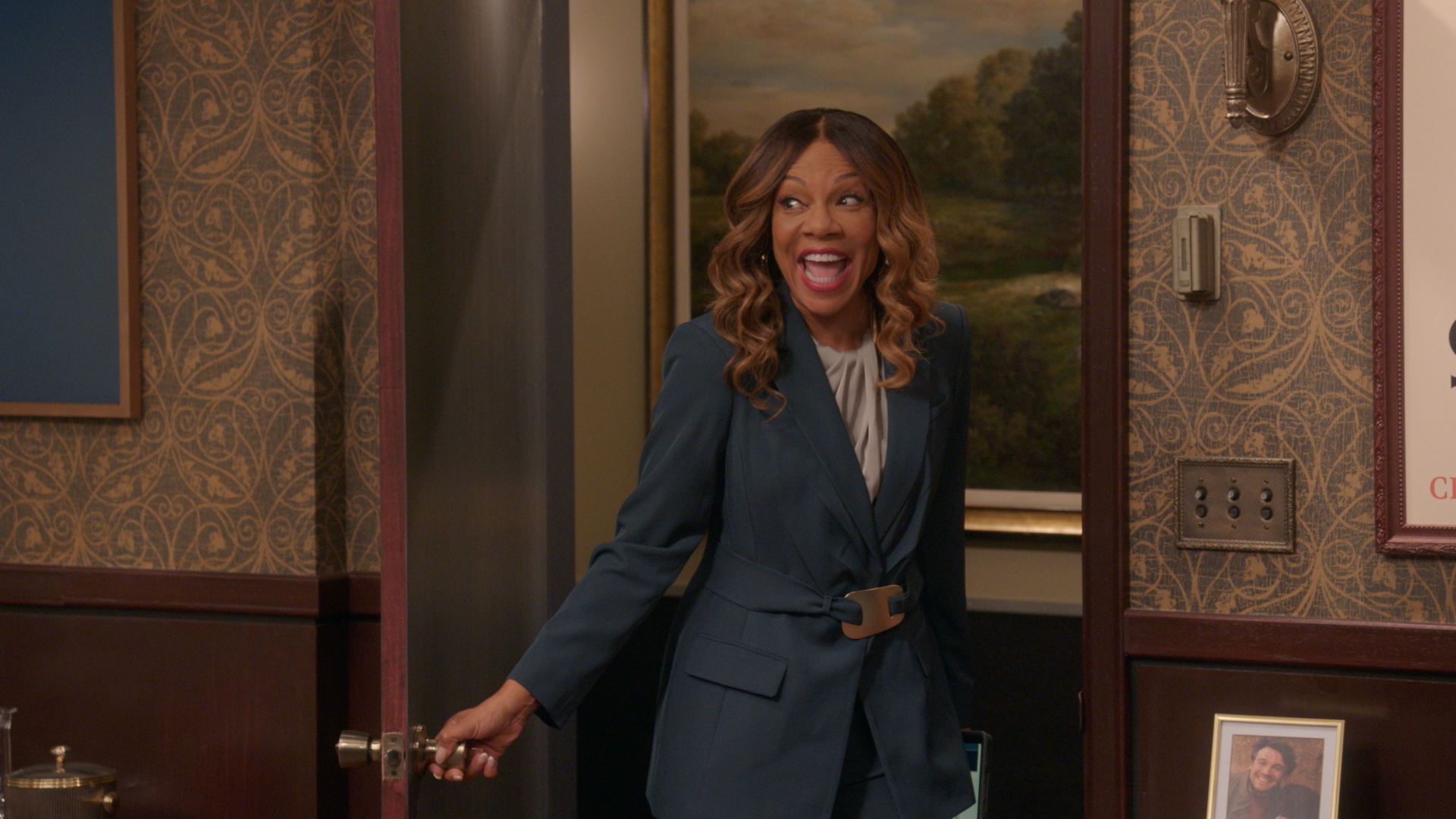 A smiling woman with curly brown hair wearing a navy blue blazer stands in a room with ornate wallpaper and framed artwork.