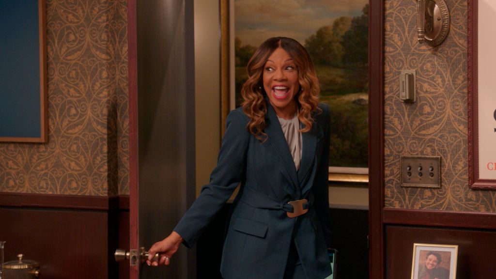 A smiling woman with curly brown hair wearing a navy blue blazer stands in a room with ornate wallpaper and framed artwork.