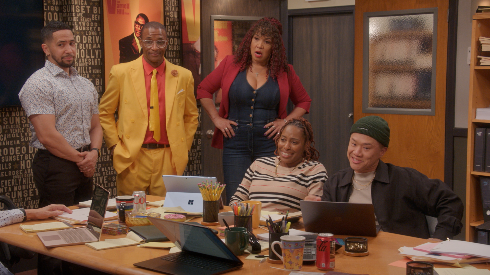 A group of five people, including two men in colorful suits, a woman in a red jacket, and two others, gathered in an office setting with various props and decor.
