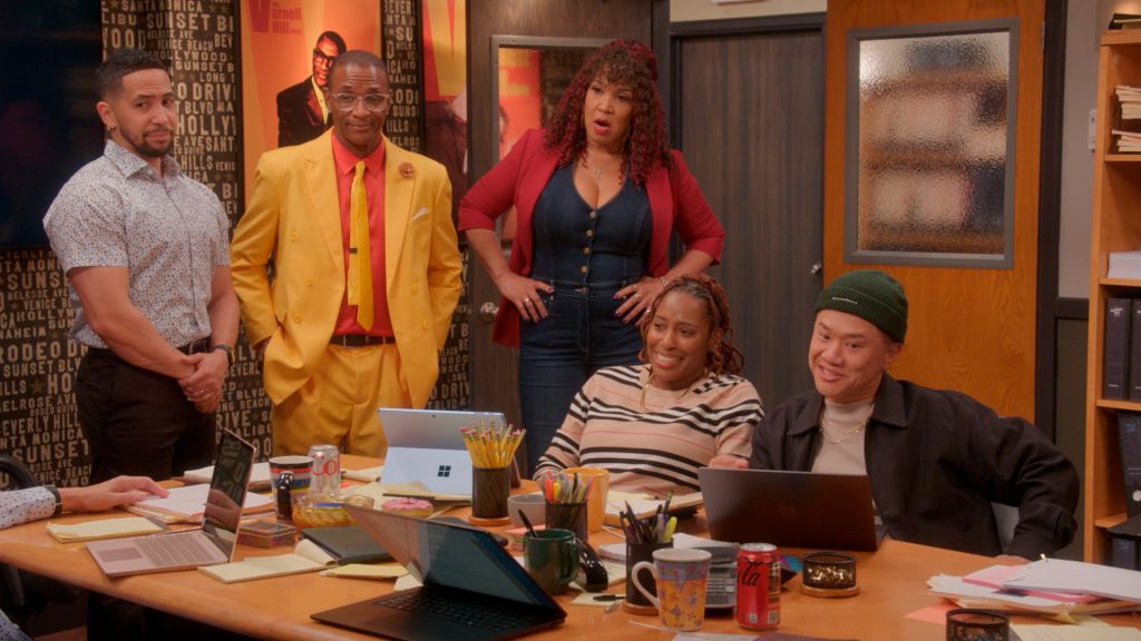 A group of five people, including two men in colorful suits, a woman in a red jacket, and two others, gathered in an office setting with various props and decor.