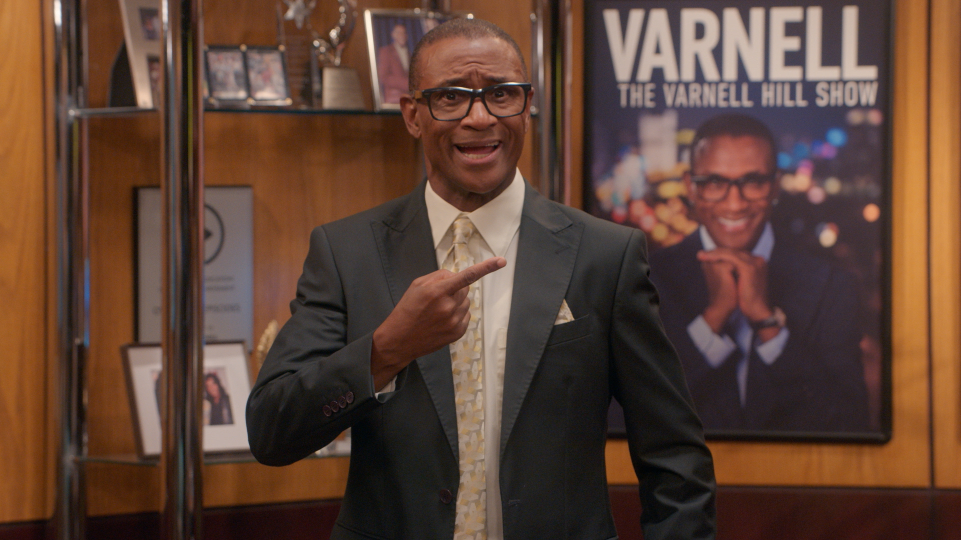A smiling man wearing glasses and a suit stands in front of a poster for "The Varnell Hill Show" on Paramount+.