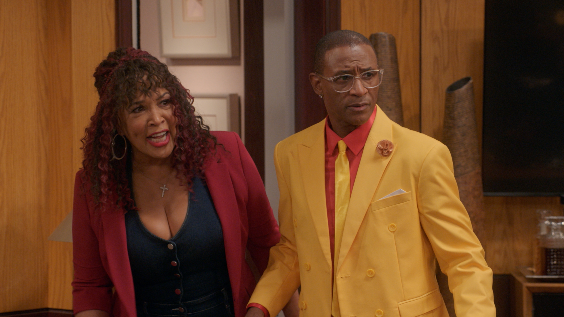 Two people, a woman with curly red hair wearing a red jacket and a man wearing a yellow suit, standing together in a room with wooden walls.