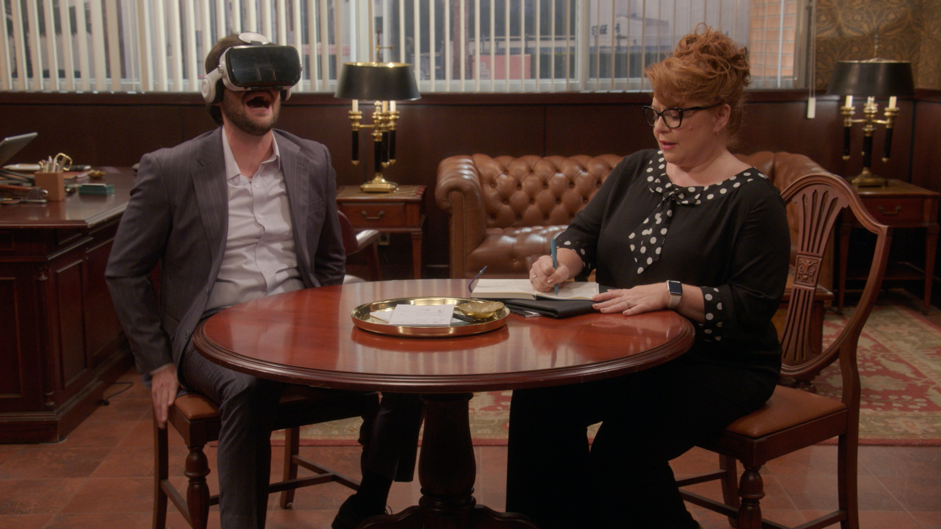 Two people seated at a table in an office-like setting, one wearing a virtual reality headset and the other taking notes.