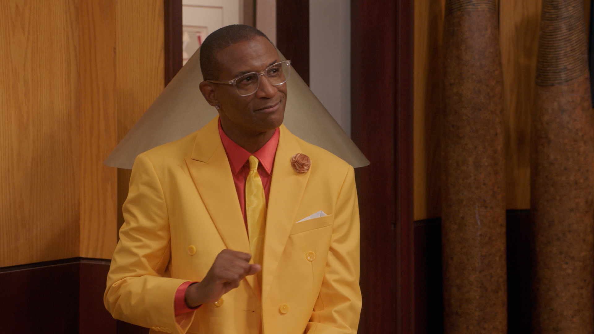 A man wearing a bright yellow suit and red tie stands in a room with wooden paneling.