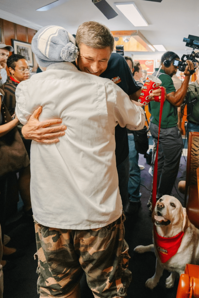A man in camouflage pants and a red jacket embracing another person in a white coat, with a dog in a red vest in the foreground.