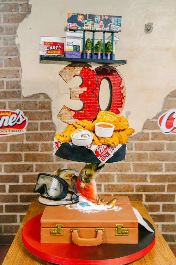 A 30th anniversary celebration display for Cane's Chicken Fingers, featuring a large red "30" number, fried chicken, dipping sauces, and a vintage-style suitcase.