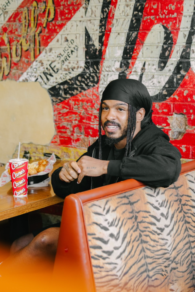 A person with dreadlocks sitting at a table, surrounded by Canes Canes Canes cups and a colorful graffiti-covered wall.
