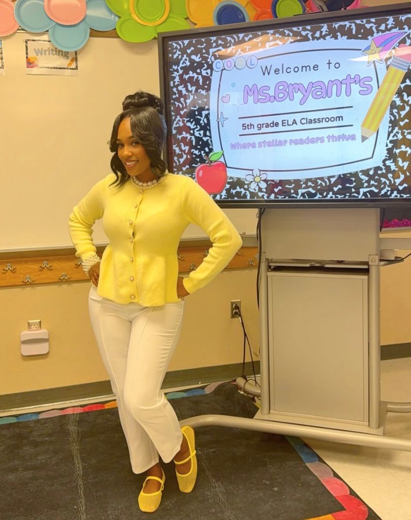 A smiling teacher wearing a yellow cardigan stands in front of a classroom welcome sign that reads "Welcome to Ms.Bryant's 5th grade ELA Classroom where stellar readers thrive".