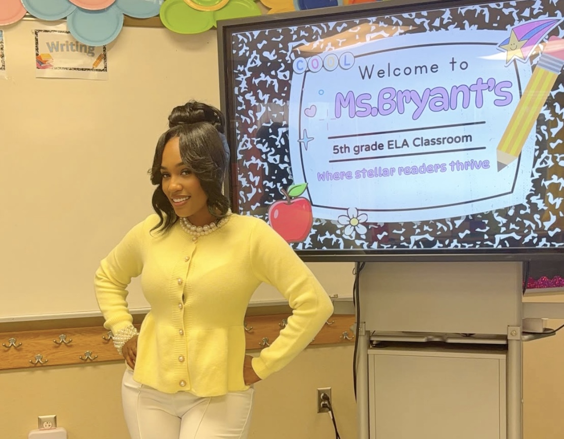A smiling woman in a yellow cardigan stands in front of a classroom welcome sign that reads "Welcome to MS.BRYANT'S 5th grade ELA Classroom where stellar readers thrive".