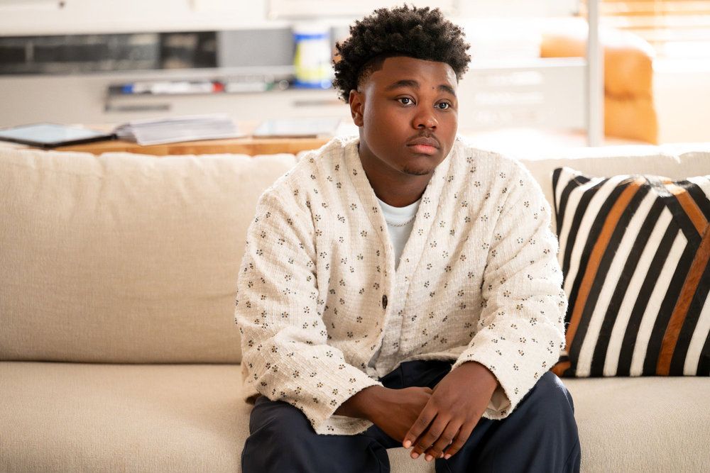 A young Black man with curly hair sits on a couch, wearing a patterned white jacket and looking thoughtfully at the camera.