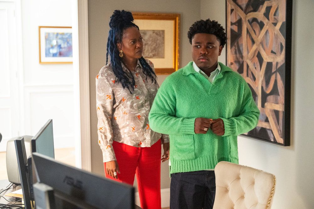 Two people, a woman with dreadlocks and a man in a green sweater, standing in a room with artwork on the walls.
