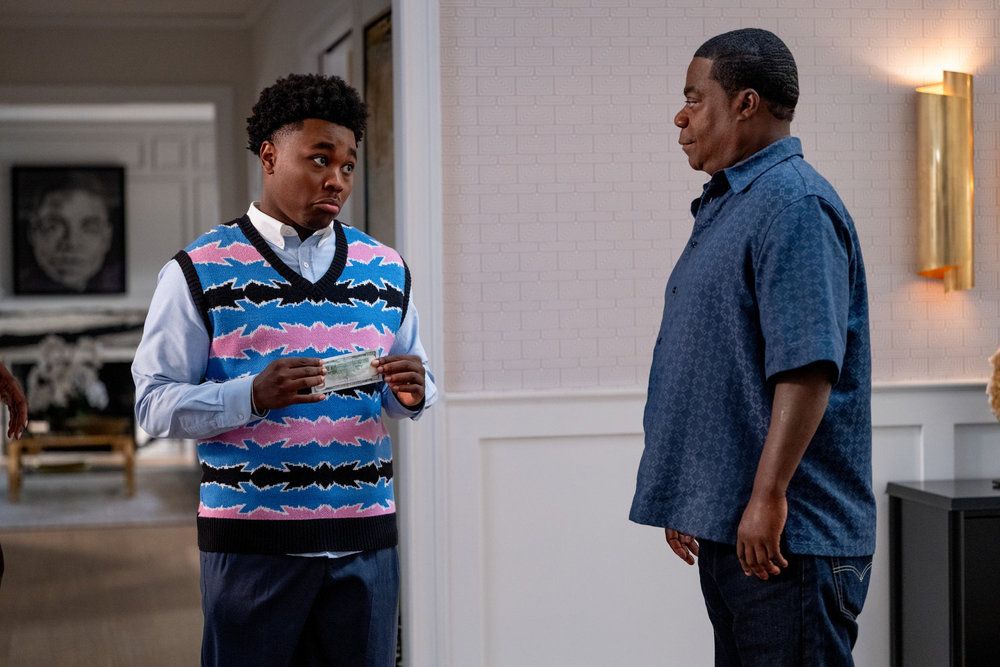 Two men, one wearing a patterned sweater vest and the other a denim shirt, standing in a room with framed artwork on the wall.