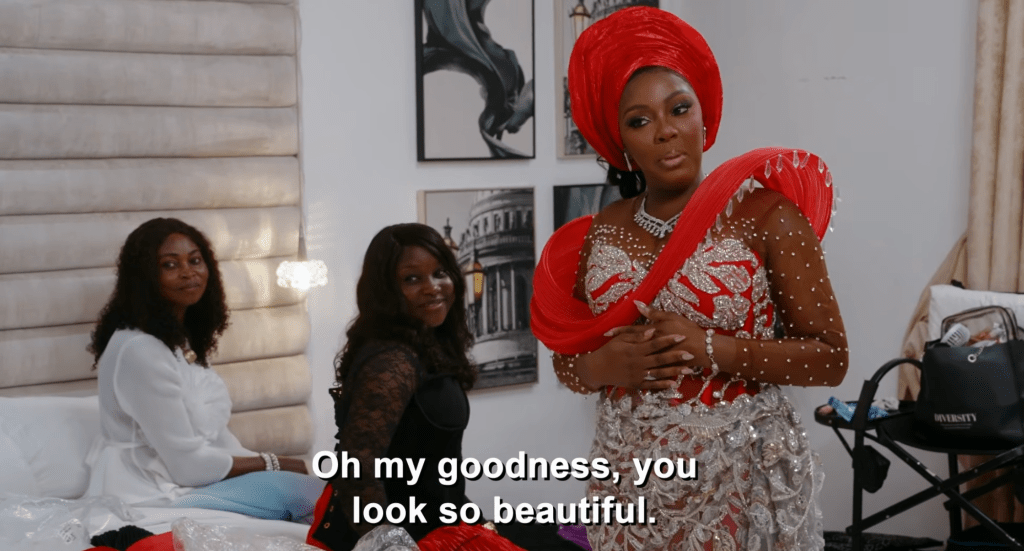 ’90 Day Fiancé’ Exclusive: Clip Ashia Sparkles & Shines In Ravishing Red For Her Nigerian Wedding–‘You Look Like A Barbie, Baby’ 6 A woman in a red dress and headpiece stands in a room with two other women, one in a white blouse and the other in a black dress. The text