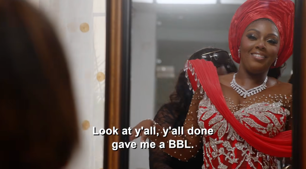 ’90 Day Fiancé’ Exclusive: Clip Ashia Sparkles & Shines In Ravishing Red For Her Nigerian Wedding–‘You Look Like A Barbie, Baby’ 4 A smiling woman wearing a red headwrap and ornate red dress with jewelry. The text overlay says "Look at y'all, y'all done gave me a BBL.